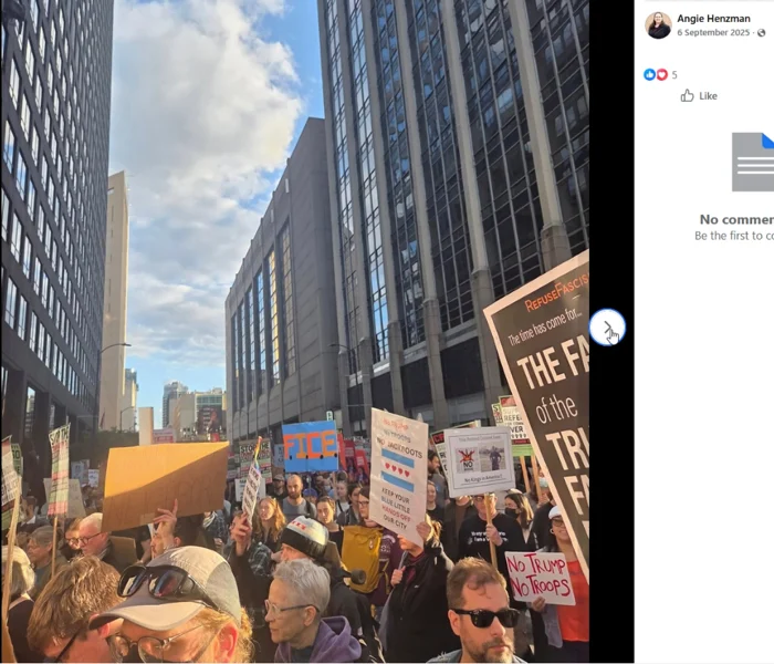 Facebook viewer screenshot of a dense protest crowd between tall downtown buildings, with placards including "No Trump No Troops" and "No Trump No Troops No Jackboots."